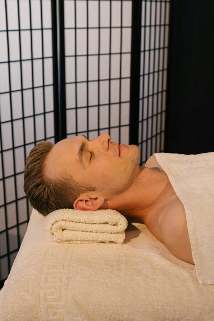 Man in a relaxing spa setting with eyes closed, lying shirtless on a towel.